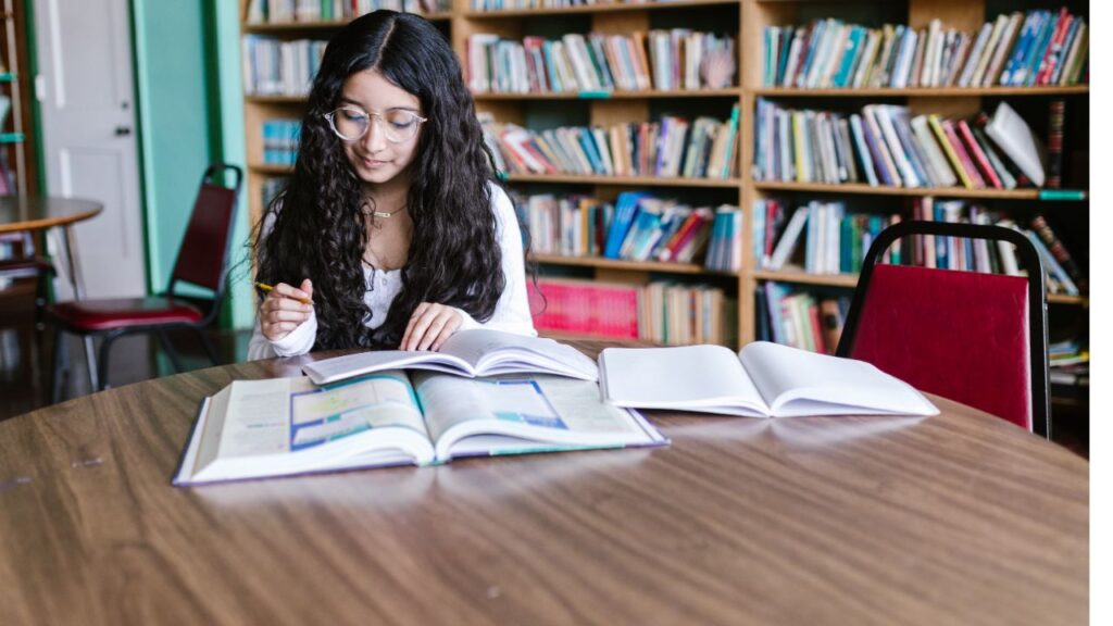 A young woman sitting at a table surrounded by books and a laptop, engaged in Writing book reviews.